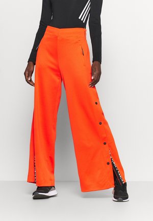 Person iført lyse orange vidde ben bukser med sorte trykknapper på siderne, sorte sneakers og en sort langærmet top med hvide striber.