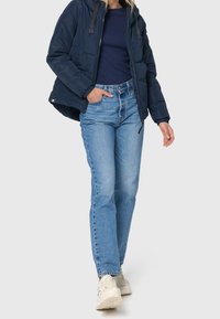 Marineblå puffjakke med hætte, parret med lyseblå højtaljede jeans. Modellen har hvide sneakers på, hænderne i lommerne. Glat stof, afslappet pasform.