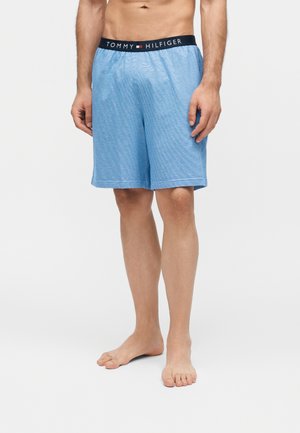 Modèle masculin portant un short bleu clair Tommy Hilfiger avec une ceinture élastique de marque, debout pieds nus sur un fond neutre.