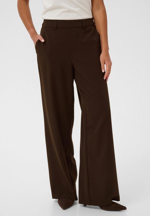 Femme portant un pantalon large marron foncé et des chaussures marron pointues, une main dans la poche et l'autre reposant à ses côtés.