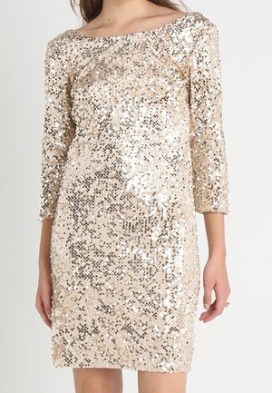 Kobieta ubrana w sukienkę sięgającą do kolan, z długim rękawem, pokrytą złotymi cekinami, z szerokim dekoltem w łódkę na jednolitym białym tle.