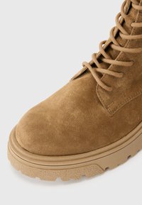 Tan suede enkelboot met vetersluiting aan de voorkant, verstevigde neus en een getextureerde rubberen zool met een uitgesproken profiel.
