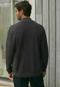 Dunkelgrauer Strickcardigan, strukturierter Stoff, Kragen-Design, lange Ärmel, gerippter Saum, über dunkel gefärbte Hosen getragen, von hinten betrachtet.