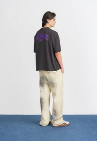 Camiseta negra de gran tamaño con el logo morado de "REEBOK" en la espalda, combinada con pantalones crema que presentan un ligero degradado y un sutil desgaste.
