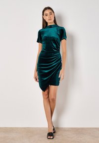 Türkisfarbenes Samtkleid mit kurzen Ärmeln, hochgeschlossenem Ausschnitt und gerafften Details an der Taille. Der Saum hat einen diagonalen Schnitt.
