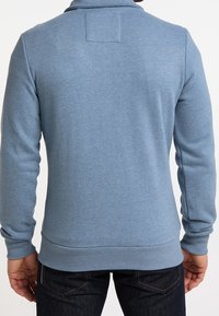 Ljusblå sweatshirt med en slät textur, med hög krage och ribbad mudd. Baksidan har en fyrkantig patchdetalj.