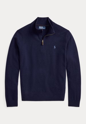 Jeune homme debout, mains dans les poches, portant un pull bleu marine, une chemise bleue, un pantalon sombre et des mocassins marron à pampilles sur fond beige.