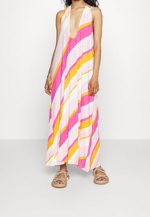 Vrouw draagt een mouwloze, kleurrijke gestreepte maxi-jurk met diepe V-hals en beige sandalen met touw, staand tegen een effen achtergrond.