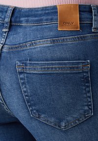 Gros plan de la poche arrière d'un jean en denim bleu avec un patch en cuir marron marqué ONLY, porté avec un haut côtelé rose.
