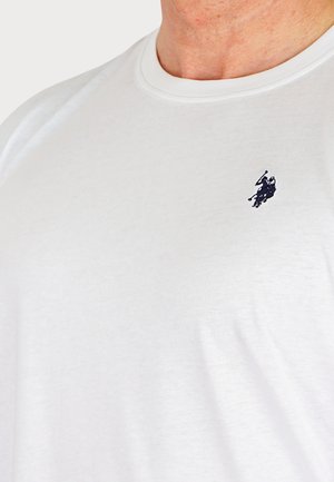 Prim-plan cu o persoană care poartă un tricou alb cu guler rotund și un mic logo negru cu jucător de polo pe partea stângă a pieptului.