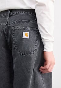 Pantalones de mezclilla gris con un bolsillo trasero que cuenta con una etiqueta blanca cuadrada y un logotipo amarillo. Una camiseta blanca de manga larga parcialmente visible.