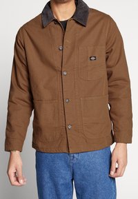 Brown cotton jacket with a corduroy collar, featuring two front pockets, button closure, and a small patch logo on the chest pocket.