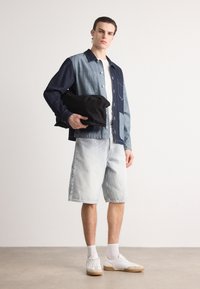 Jachetă din denim cu panouri albastre deschise și închise, tricou alb, pantaloni scurți albaștri deschisi, ținând o geantă neagră. Încălțăminte albă cu tălpi bej.