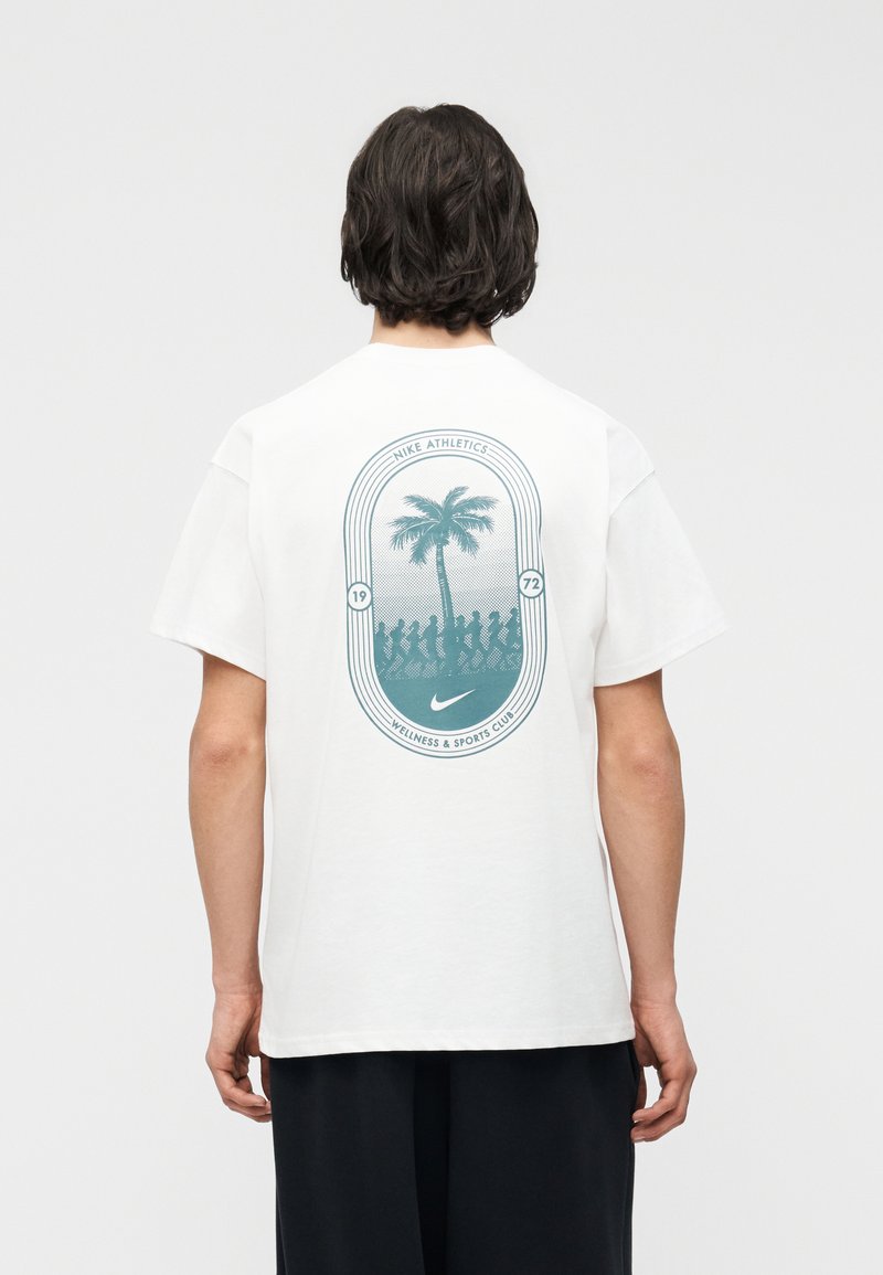 Άτομο με σκούρα μαλλιά που φοράει λευκό T-shirt με τυρκουάζ γραφικό από φοίνικα, φιγούρες που τρέχουν και κείμενο "Nike Athletics Wellness & Sports Club".