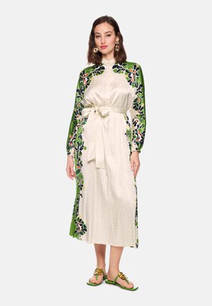 Woman wearing a long cream dress with green floral-patterned sleeves and sides, tied at the waist, paired with green sandals and earrings.