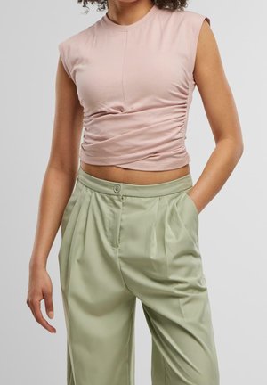 Vrouw draagt een mouwloze lichtroze gerimpelde cropped top en losse, lichtgroene, hoge taillebroeken met één hand in de zak.