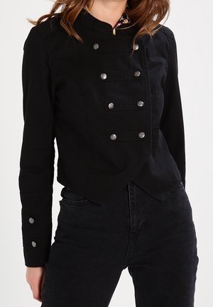 Frau trägt eine taillierte schwarze Militärjacke mit silbernen Knöpfen und eine hoch taillierte schwarze Jeans.