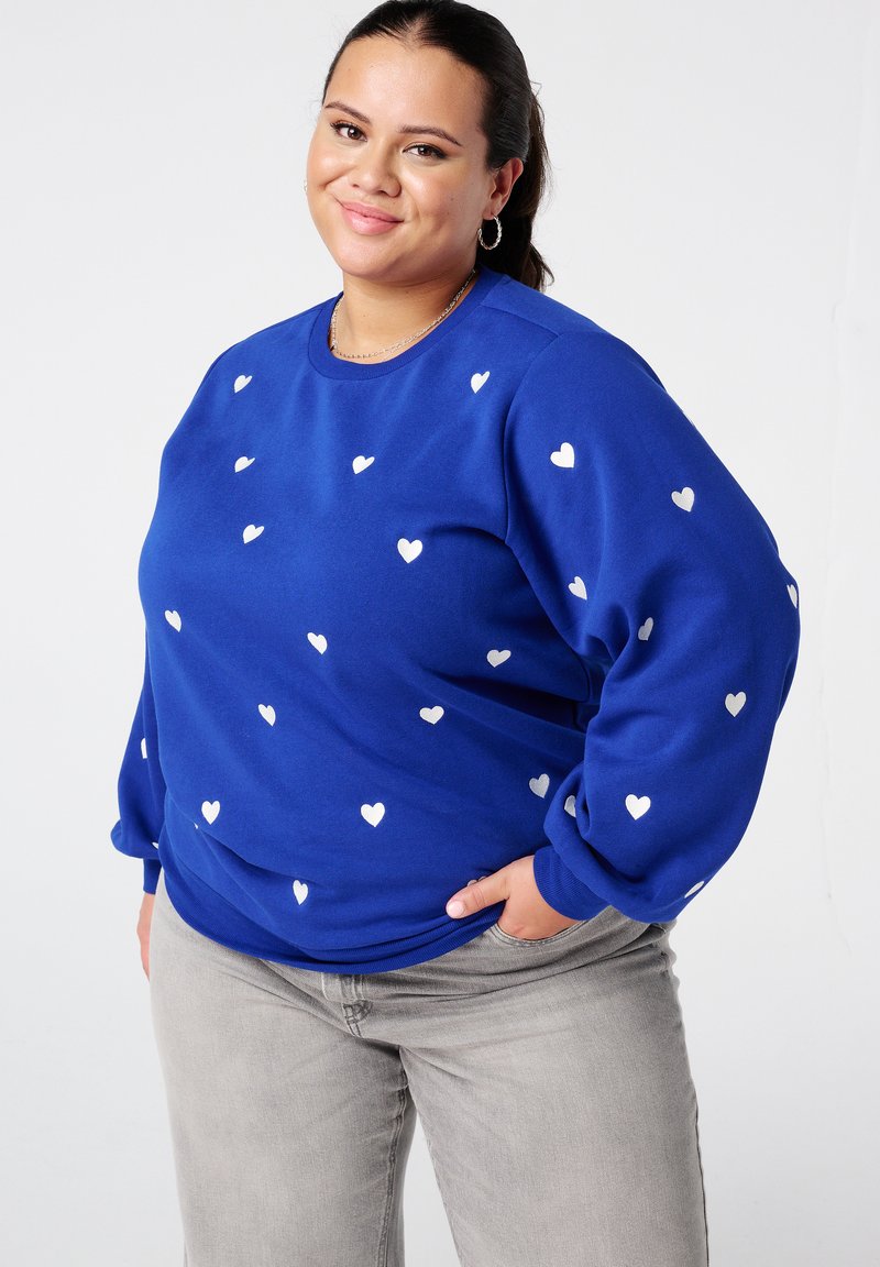 Sweat-shirt bleu avec motifs de cœurs blancs, col rond et manches longues. Associé à un jean gris clair. Texture de tissu douce visible.