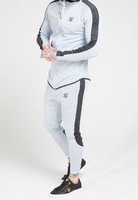 Lyseblå atletisk træningsdragt med sorte detaljer, bestående af en jakke med lynlås og tætsiddende bukser. Materialet er mesh og har et struktureret mønster.