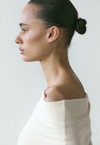 Widok z profilu modelki ubrane w jasnobeżową bluzkę odkrywającą ramiona, o gładkiej fakturze i czystych liniach na jednolitym tle.