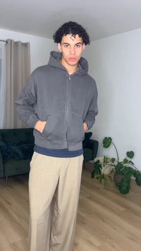 Tânăr cu păr creț, purtând un hanorac gri cu fermoar și pantaloni bej, stă într-un living modern cu plante și o canapea verde.