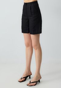 Pantaloni corti neri in lino con una vestibilità sartoriale, dotati di pieghe frontali e tasche laterali. Il materiale presenta una leggera trama e un aspetto leggero.