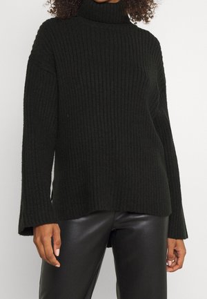 Femme portant un pull à col roulé côtelé noir et un pantalon en cuir noir, debout devant un fond blanc uni.