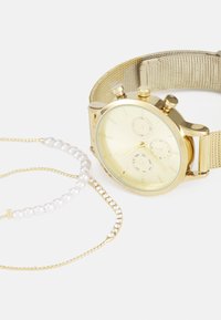 Montre-bracelet en or avec bracelet en maille à côté de deux bracelets en or délicats, l'un avec des perles et l'autre avec de petites pierres transparentes, sur fond blanc.
