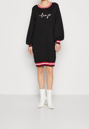 Kobieta w czarnej sukience z długim rękawem z czerwonym i różowym wykończeniem, z napisem „liu jo” na piersi, w białych botkach, stojąca na jednolitym tle.