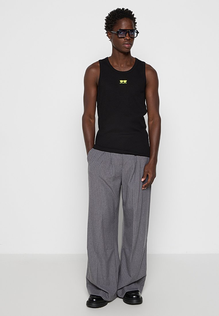 Hombre con una camiseta sin mangas negra con un pequeño logo amarillo, pantalones de rayas grises de pierna ancha, zapatos negros y grandes gafas de sol oscuras, apoyado contra una pared blanca.