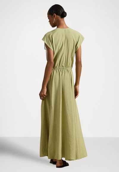Γυναίκα με αμάνικο μάξι φόρεμα ελιάς με ζωνάρι στη μέση, στέκεται ξυπόλυτη σε απλό φωτεινό φόντο, με την πλάτη γυρισμένη.