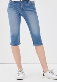Pantaloni capri in denim azzurro chiaro con un design aderente, taglio fino al ginocchio e stile a cinque tasche. Abbinati a sneakers bianche con dettagli neri.