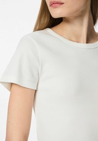 Ribota, īsa piedurkne, balta blūze ar apaļo kakla izgriezumu. Auduma struktūra ir gluda, ar smalkiem vertikāliem rakstiem. Tuva skatiena skats uz audumu un plecu.