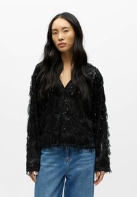 Žena s dlouhými černými vlasy má na sobě černou flitrovanou bundu přes modré džíny a stojí před jednoduchým bílým pozadím.