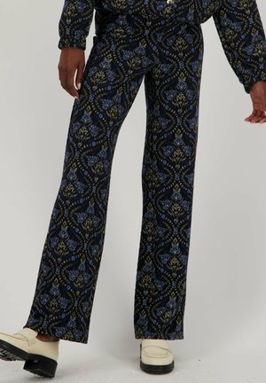 Pantalons évasés en tissu bleu marine avec un motif floral doré et bleu. Design texturé avec une finition lisse. Associés à des chaussures crème.