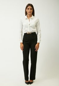 Camisa blanca de botones con adornos en el cuello, combinada con pantalones negros de cintura alta. Cinturón de cadena dorada. Tacones negros de punta. 