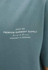 Zelená tkanina trička s bílým textem "Od roku 1990 Prémiová oděvní dodávka Jack & Jones Navrženo v Dánsku" výrazně zobrazeným.