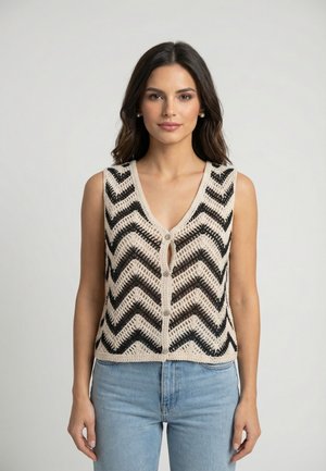 Kobieta o ciemnych włosach, nosząca perłowe kolczyki, bezrękawnik w kolorze beżowo-czarnym w jodełkę i jasnoniebieskie dżinsy, stojąca na jednolitym tle.