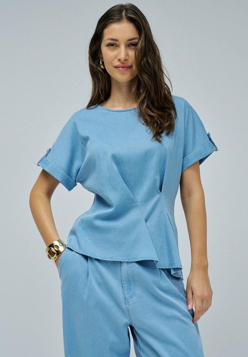 Top in denim blu chiaro con maniche corte arrotolate, scollatura rotonda e orlo asimmetrico, che presenta un tessuto texturizzato e un design minimalistico.