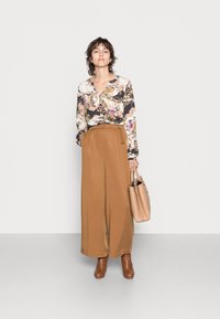 Blusa floral com mangas longas e design envolvente, combinada com calças largas de cintura alta em tom castanho. Inclui uma bolsa de mão em tom de pele clara e botas castanhas.