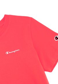 Koralfarvet t-shirt lavet af bomuld; har en rund halsudskæring, korte ærmer og hvidt "Champion" logo på venstre bryst og ærme.