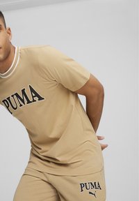 Camiseta de manga corta beige con el logo negro de "PUMA" y un cuello blanco acanalado, combinada con pantalones cortos beige a juego. Material de algodón suave.