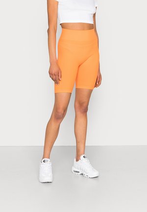 Osoba ubrana w wysokie, jaskrawo pomarańczowe, krótkie spodenki rowerowe, biały krótki top oraz białe sneakersy Nike Air Max stojąca na jasnoszarej podłodze.