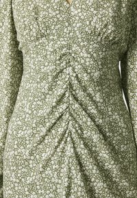 Grünes Blumenkleid mit weißen Mustern, gerafften Details, langen Ärmeln und V-Ausschnitt, aus weichem Stoff mit strukturierter Oberfläche gefertigt.