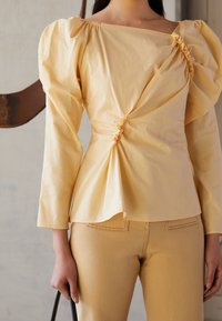 Kvinna i en ljusgul topp med rynkade detaljer och puffiga axlar, i kombination med matchande högmidjade byxor, som håller i handtaget på en svart väska.