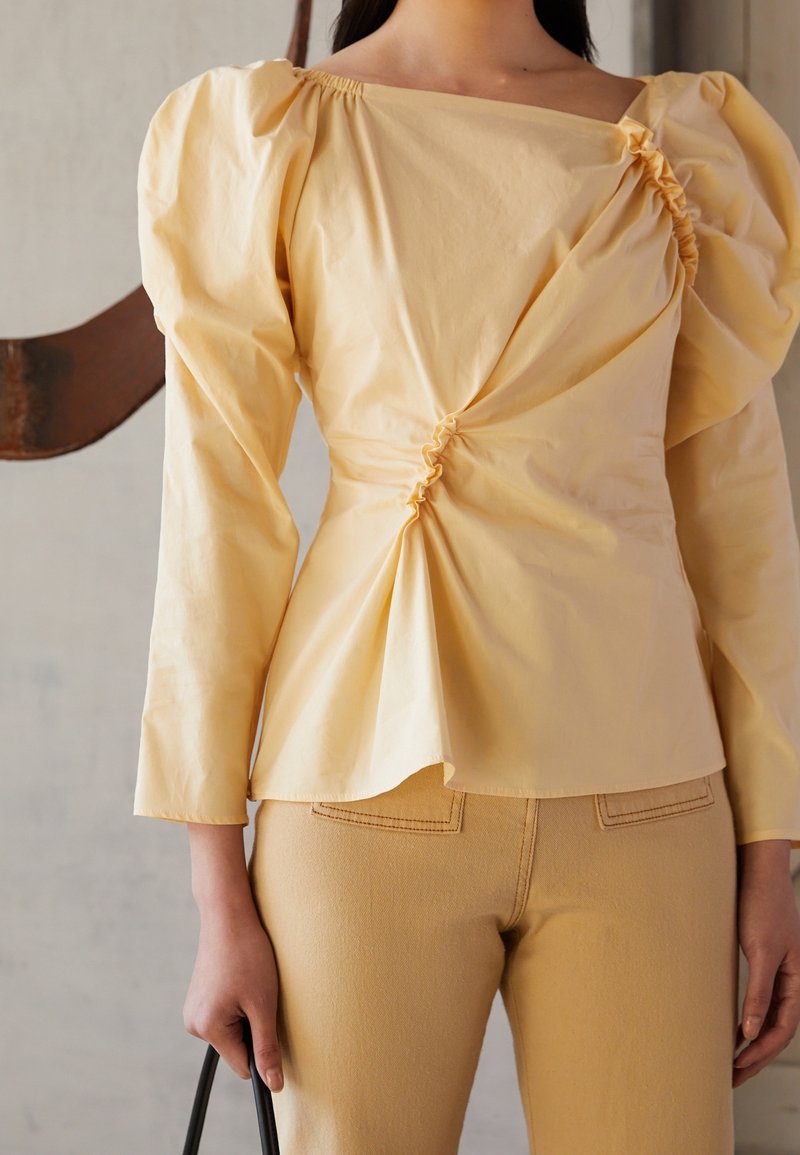 Kvinna i en ljusgul topp med rynkade detaljer och puffiga axlar, i kombination med matchande högmidjade byxor, som håller i handtaget på en svart väska.