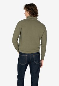 Suéter verde de punto con cuello alto, puños acanalados y cintura; combinado con jeans azul oscuro, visto desde la parte de atrás.