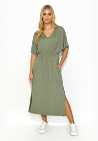 Oliwkowa maxi sukienka z miękkiego materiału, z dekoltem w serek, krótkimi rękawami, elastyczną talią z detalem guzikowym oraz bocznymi rozcięciami, zestawiona z białymi sneakersami.