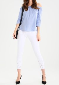 Niebieska bluzka w paski odkrywająca ramiona z rękawami do połowy przedramienia, zestawiona z białymi obcisłymi spodniami z kwiatowymi wycięciami na dole. Czarne szpilki.