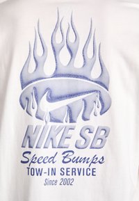 Bílý tričko s modrou grafikou zobrazující plameny a text: "NIKE SB Speed Bumps Tow-in Service Since 2002" v různých písmenech.